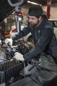 Automotive technician wearing gloves and a backwards cap working under the hood of a vehicle in a repair shop.