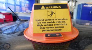Safety warning sign on a hybrid vehicle in a repair shop, cautioning about high-voltage electricity risk during service.