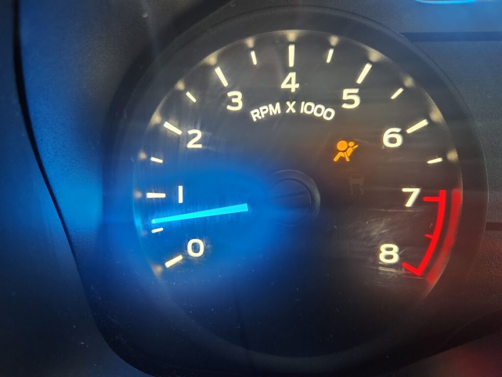 A close-up view of a vehicle's tachometer showing the RPM gauge and an illuminated amber airbag warning light. The icon, located between the 4 and 6 RPM marks, indicates a potential issue with the airbag system.