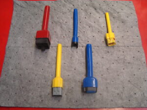 A set of five airbag connector safety tools in various colors (red, yellow, and blue) arranged on a perforated grey mat. These tools are used to safely handle airbag squib connectors and prevent accidental deployment during service.
