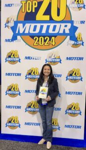 Victoria Repice at MOTOR’s booth during AAPEX 2024, smiling while holding a MOTOR Top 20 Award plaque in front of the MOTOR Top 20 branded backdrop.