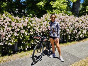 Woman smiling outdoors while standing next to a black road bike in front of a blooming wall of pink flowers on a sunny day.