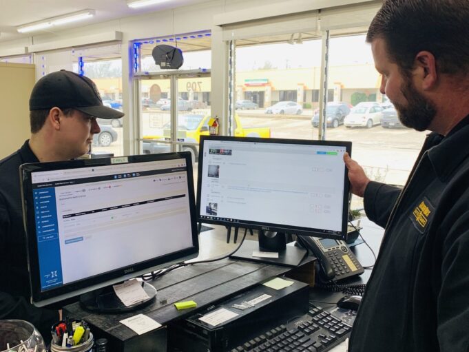 Automotive service professionals using MOTOR software on dual computer monitors at a repair shop counter to manage repair orders and customer information.