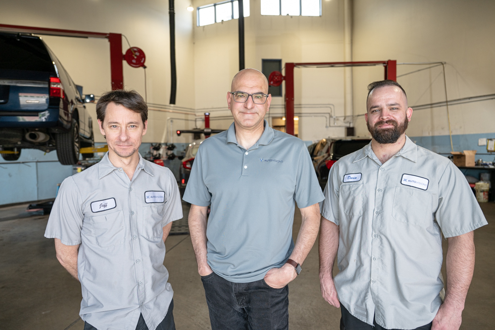 Nathan Bryant (center) with two team members standing inside the Autovisions repair shop. All three wear branded work shirts, representing a unified and personable shop team.