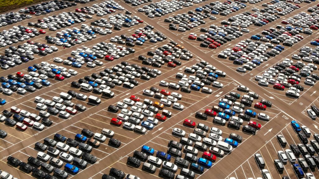 Aerial view of a massive parking lot filled with rows of new cars in various colors, representing automotive manufacturing, dealership inventory, and vehicle distribution.
