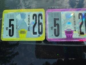 Close-up of Pennsylvania state vehicle inspection stickers on a windshield, showing a yellow-bordered 2026 annual emission sticker and a purple-bordered 2026 annual windshield sticker, both labeled "IM 5" for May expiration. The stickers display serial numbers and official state imagery under direct sunlight.
