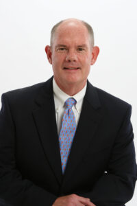 Professional headshot of a middle-aged man in a dark suit jacket, white dress shirt, and blue tie with red paisley patterns, posing against a plain white background.
