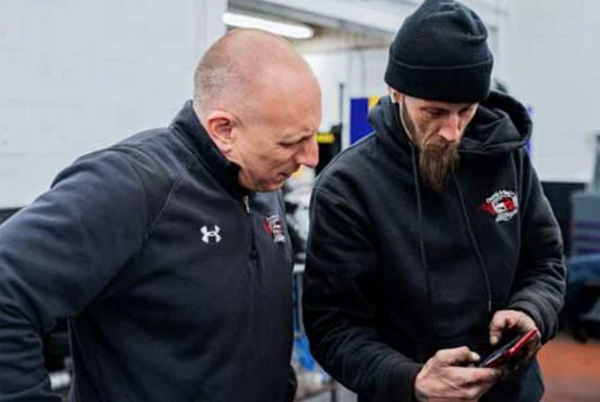 Two auto repair professionals in branded shop uniforms reviewing repair details on a mobile device inside a service bay.